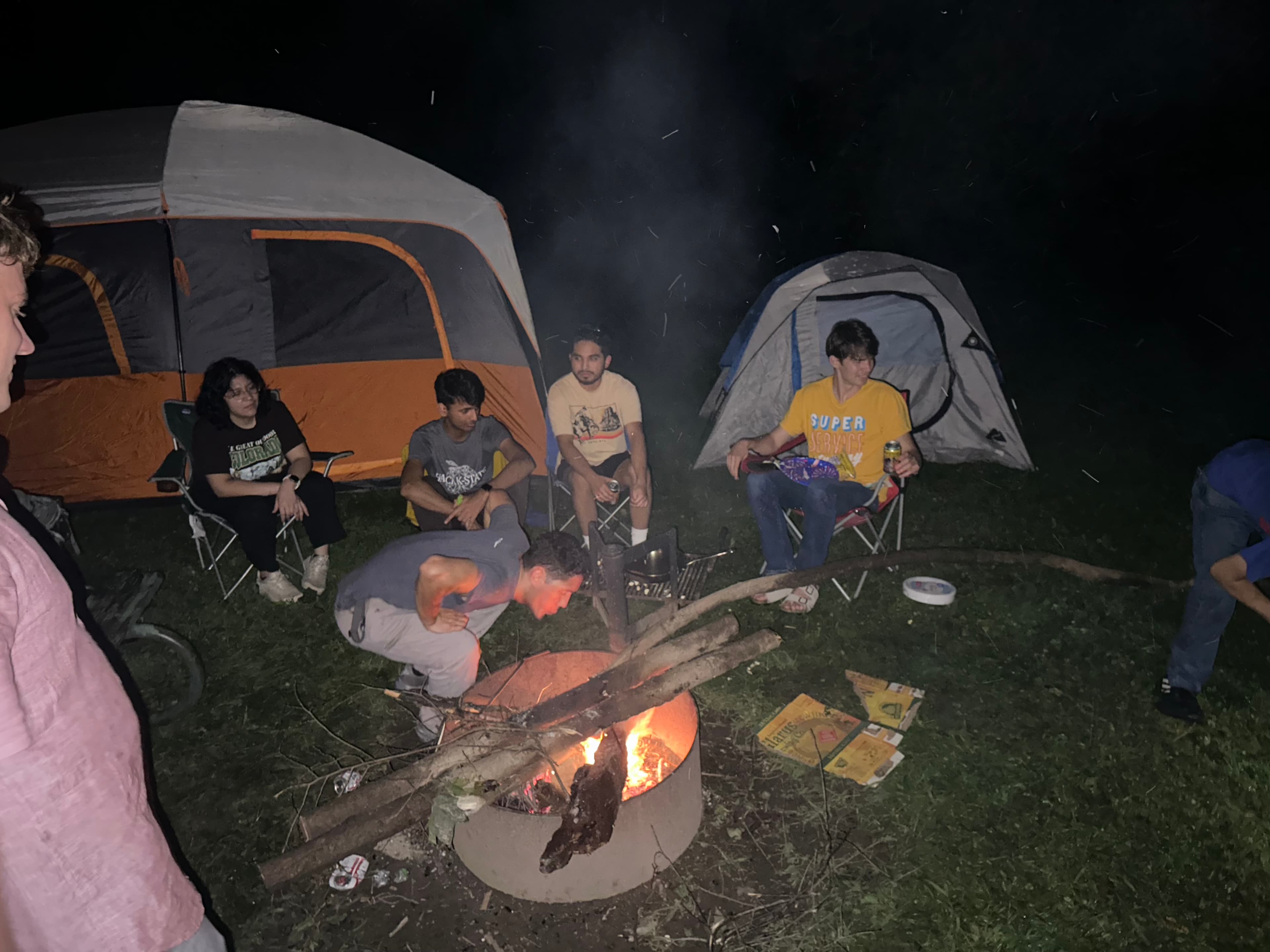 Friends in camp chairs around a fire ring at night, with two tents behind them and one person blowing on the fire