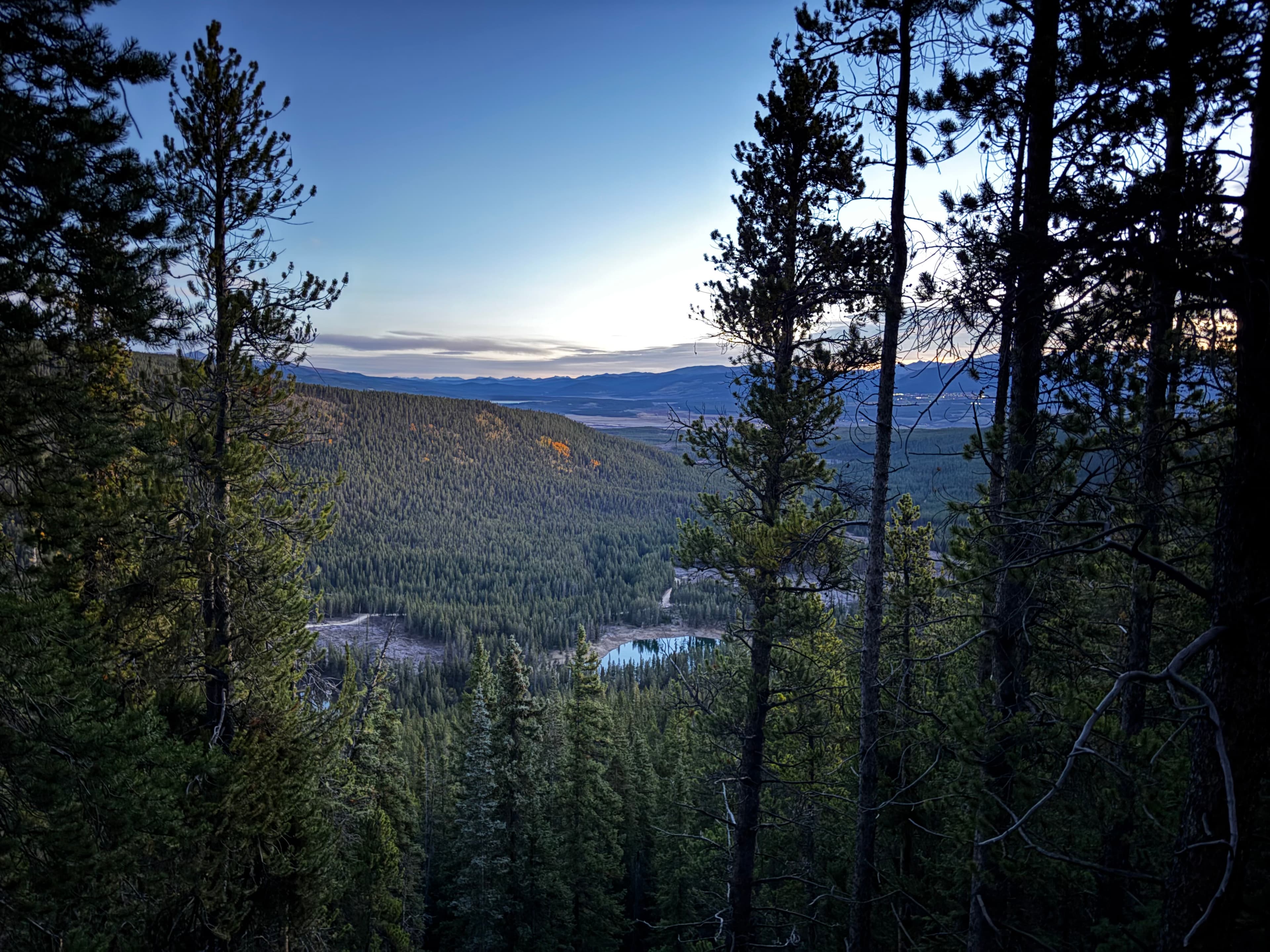 First light over a forested valley and a small lake, seen through pine trees on the way up Mount Elbert